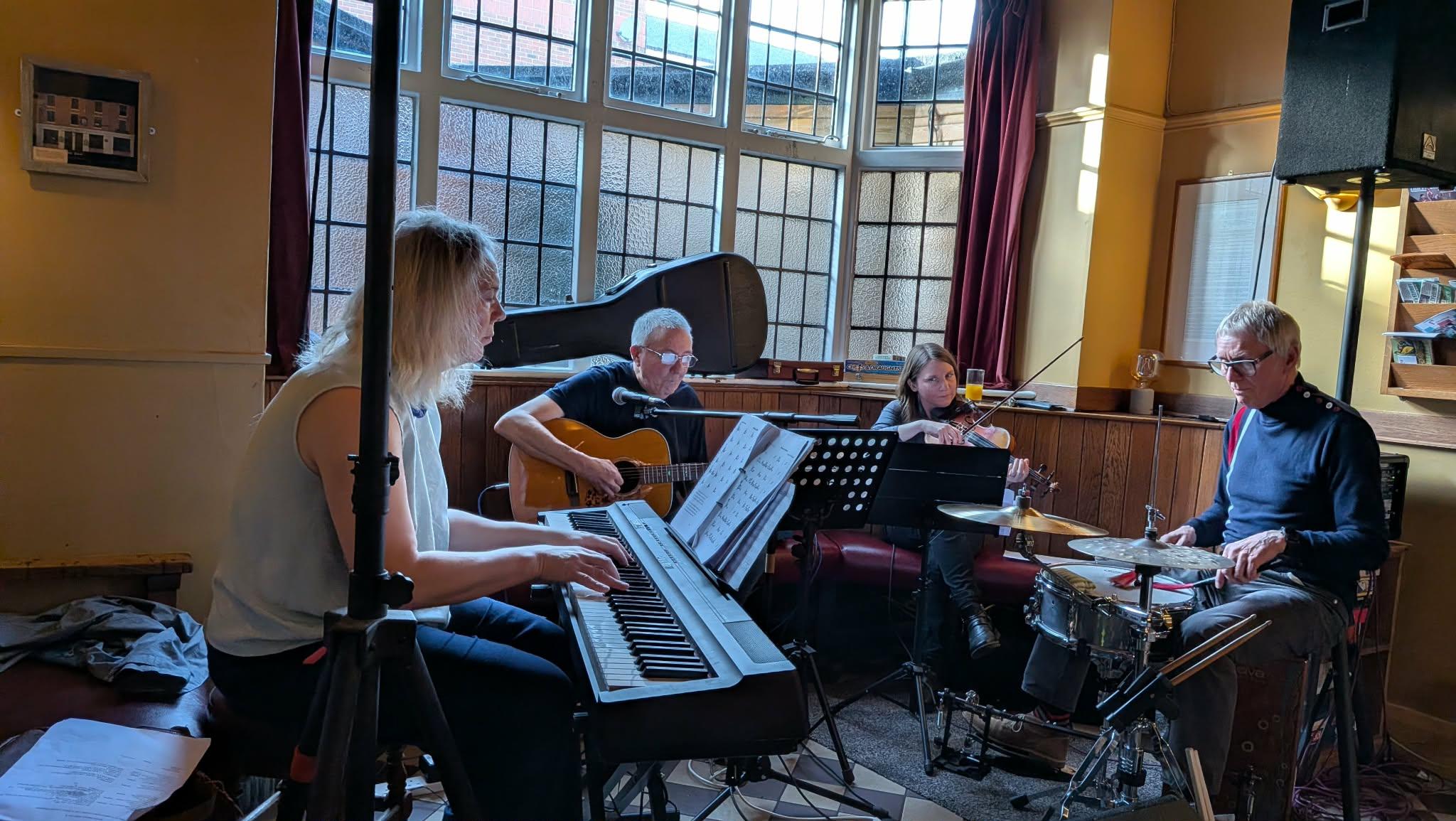 A wider photo of people playing instruments in a bar. Piano, guitar, violin, drums